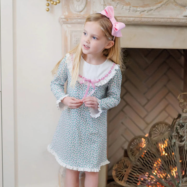 Young girl in a floral dress with a pink bow standing in front of a fireplace.