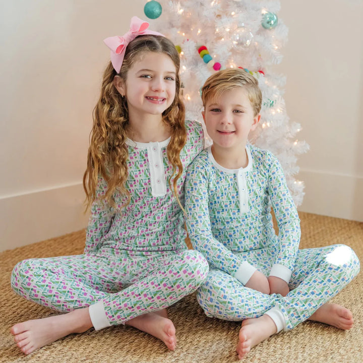 Two children in matching pajamas sitting on a carpeted floor with a decorated Christmas tree in the background.