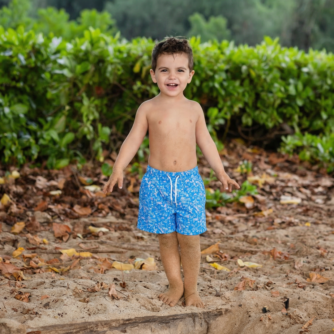 Child wearing blue swim shorts standing on a sandy path with greenery in the background