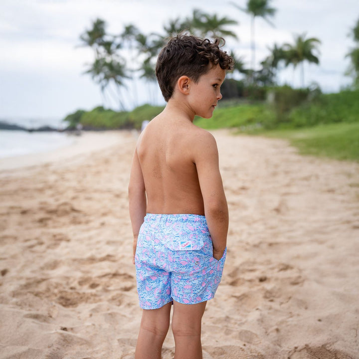 Young boy standing on a sandy beach wearing colorful swim shorts.