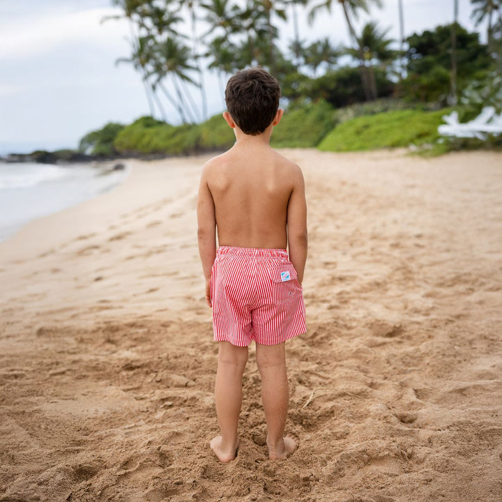Child wearing pink swim trunks on a sandy beach with palm trees in the background