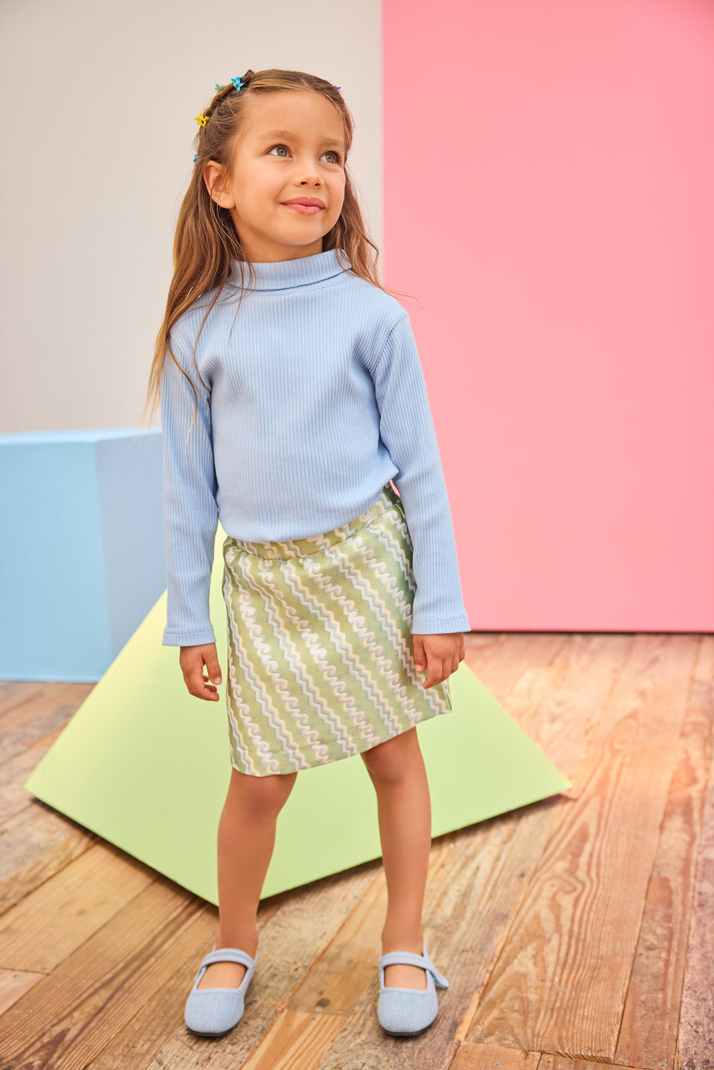 Young girl in a light blue sweater and patterned skirt standing on a wooden floor with colorful steps in the background.