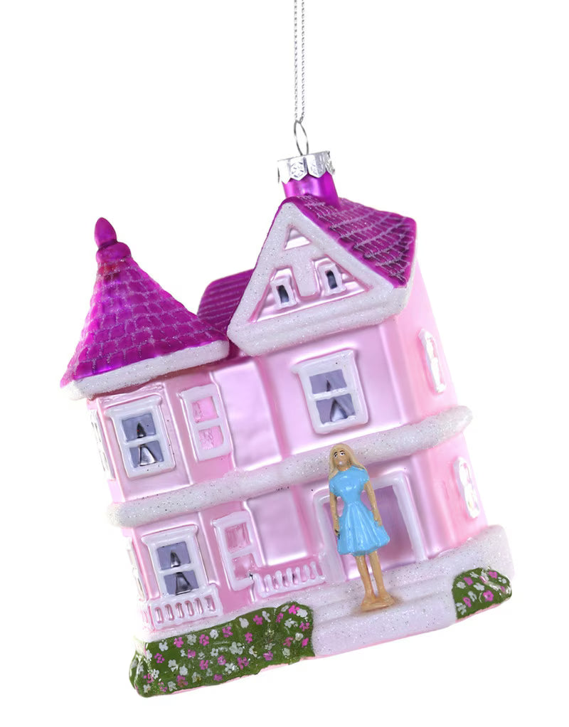 A pink and purple hanging ornament shaped like a vintage house, with a small figure of a woman in a blue dress standing on the front porch, shown against a plain white background.