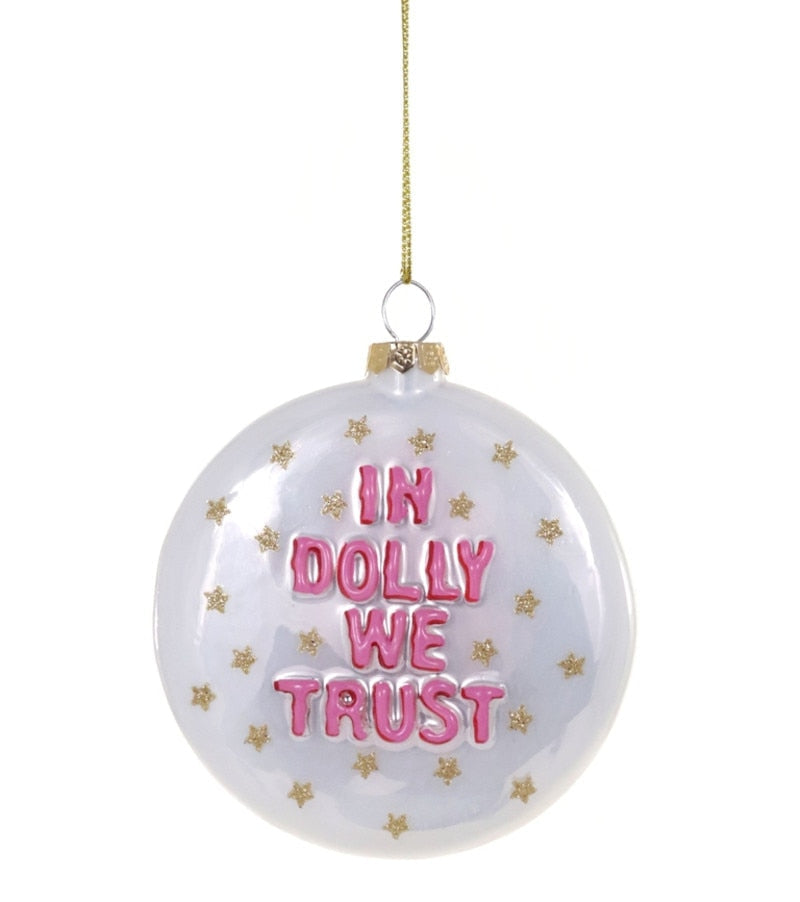 A round clear glass Christmas ornament with a gold hanging string, featuring the text 'IN DOLLY WE TRUST' in pink letters and small gold stars on a white background.