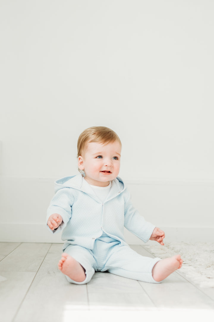 Baby wearing a light blue hoodie and pants sitting on a white floor.