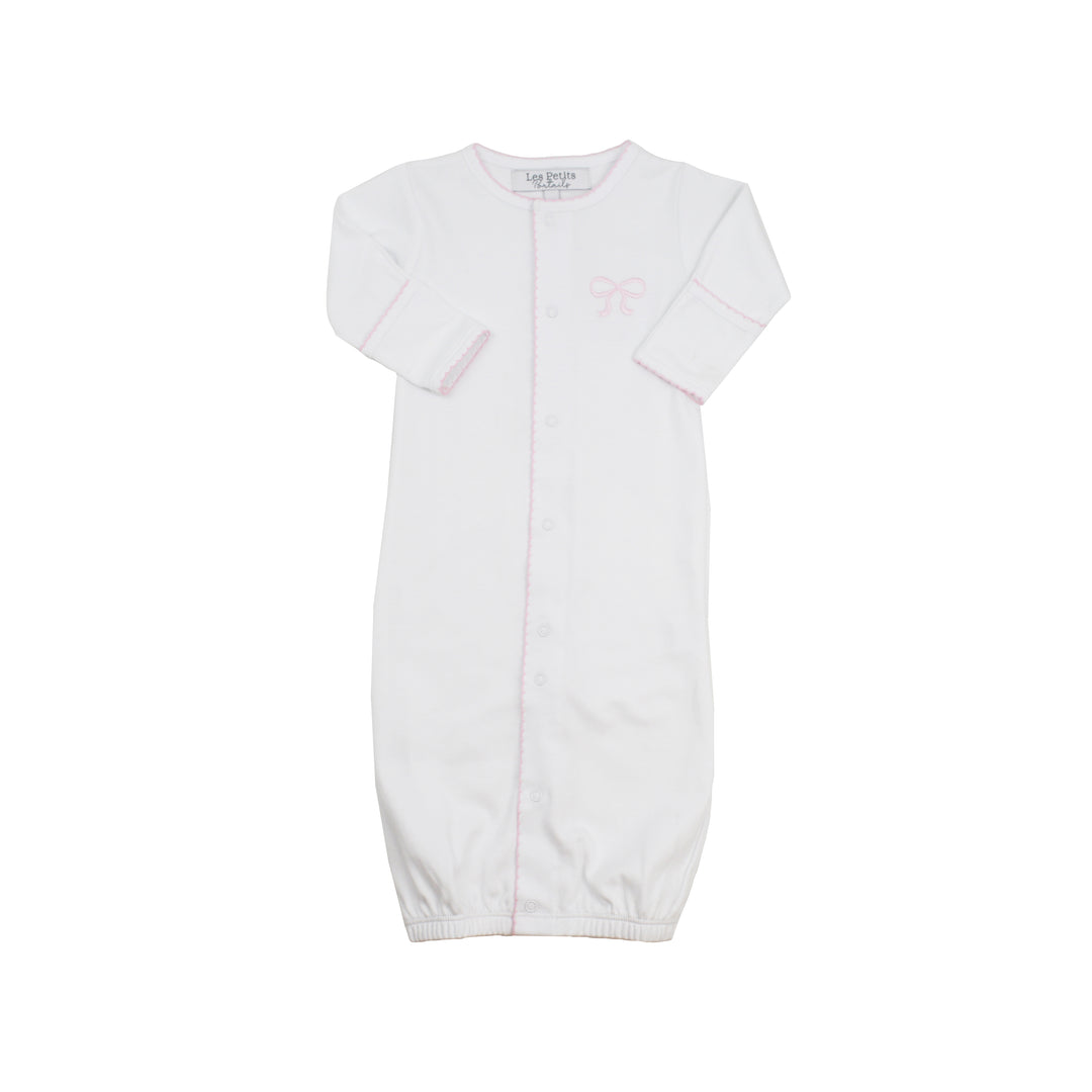 A white long-sleeve baby sleep sack with a button front, featuring a small light pink bow graphic on the left chest and a pink piping detail along the center seam.