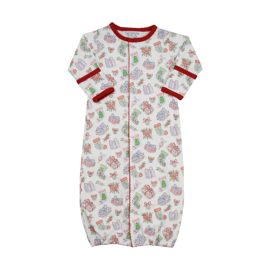 A baby sleep sack with a festive Christmas print, featuring a white background with illustrations of wrapped presents, Christmas trees, and Santa Claus. It has red trim along the collar, cuffs, and center front zipper.