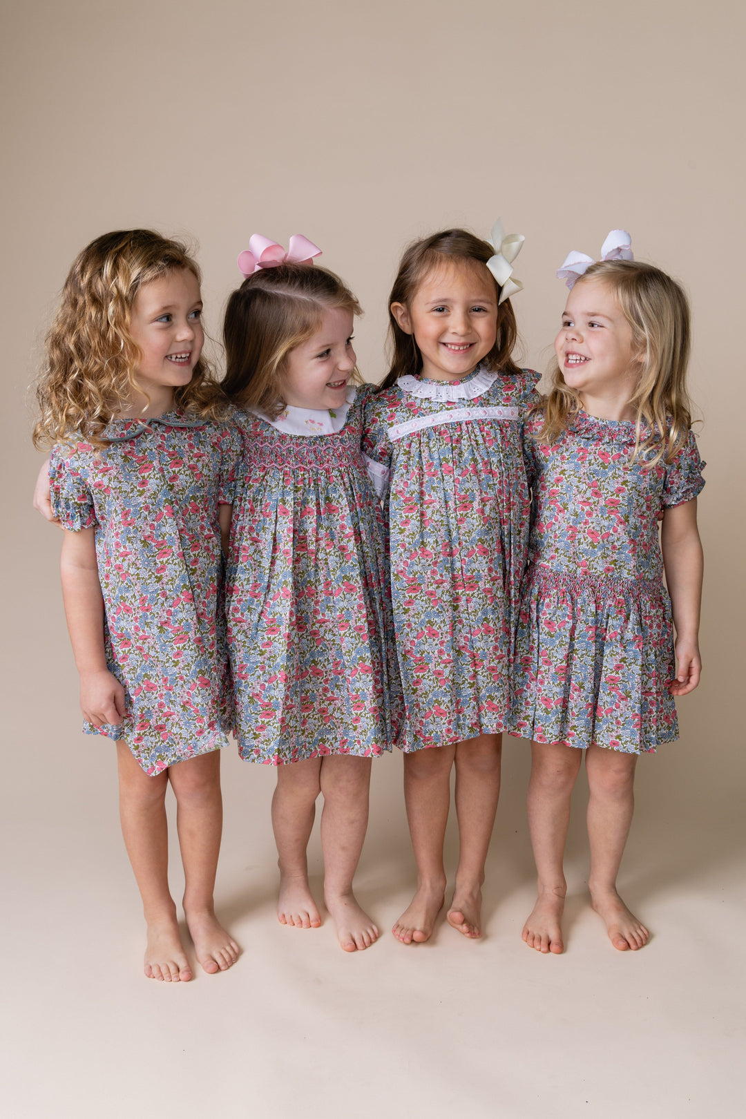 Four young girls standing barefoot against a plain beige studio background, wearing matching floral short-sleeve dresses with white ruffled collars and bow hair clips in pink, white, and lavender.