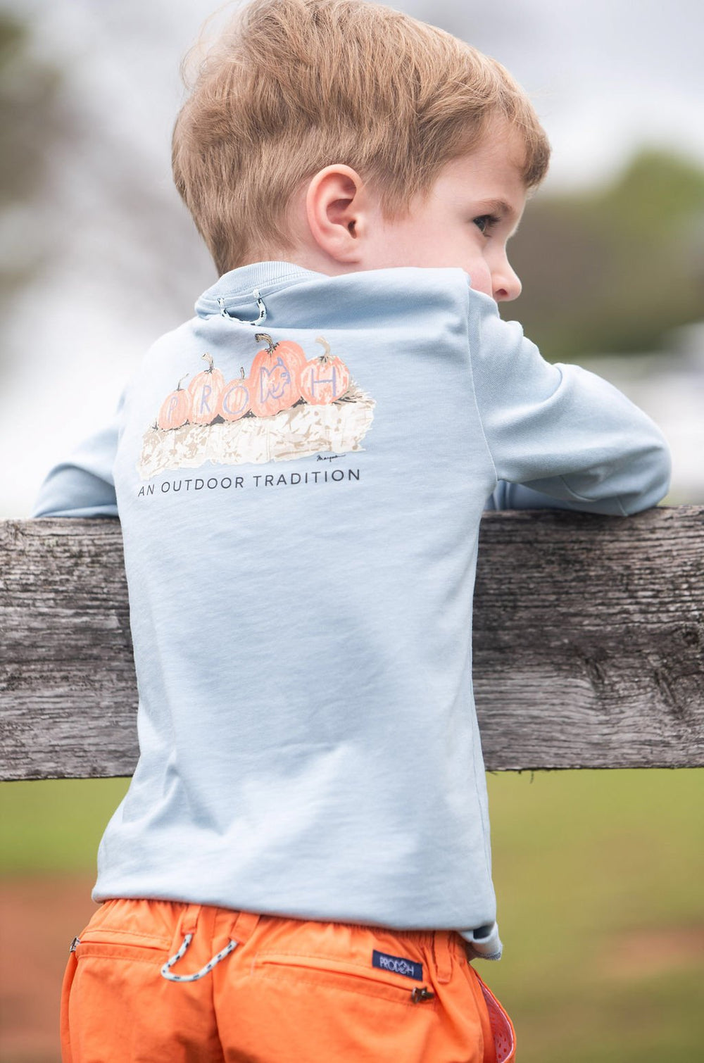 Child wearing a light blue jacket with branding, standing by a wooden fence outdoors.