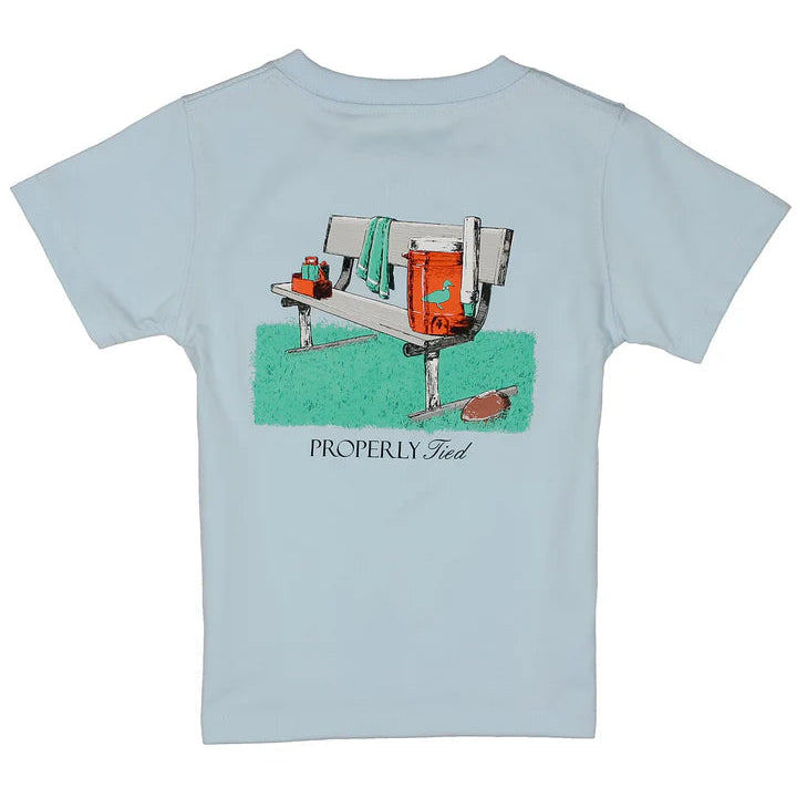 A light blue short-sleeve t-shirt with a vintage-style graphic of a bench and football cooler, featuring the text &