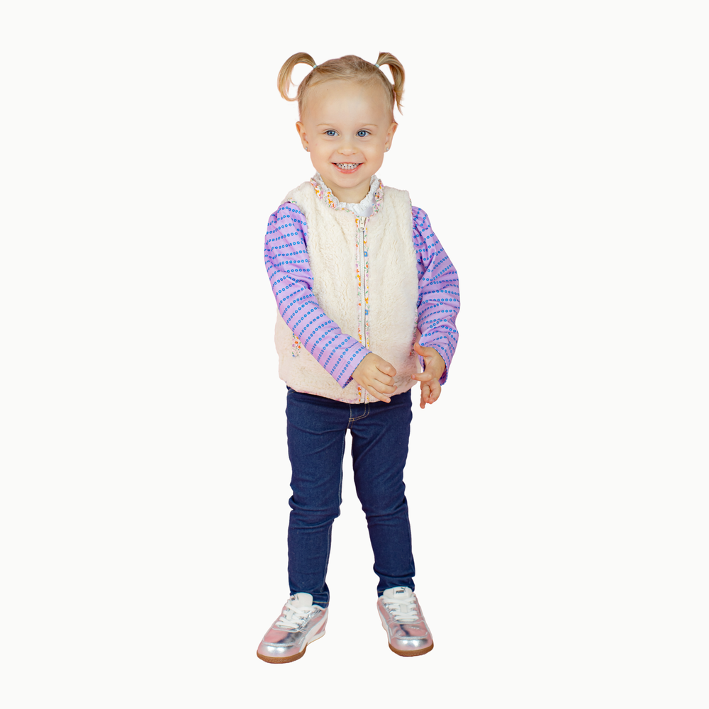 A young child with blonde pigtails stands against a plain white background wearing a cream-colored vest over a purple and white patterned long-sleeve shirt, dark blue jeans, and metallic silver sneakers.