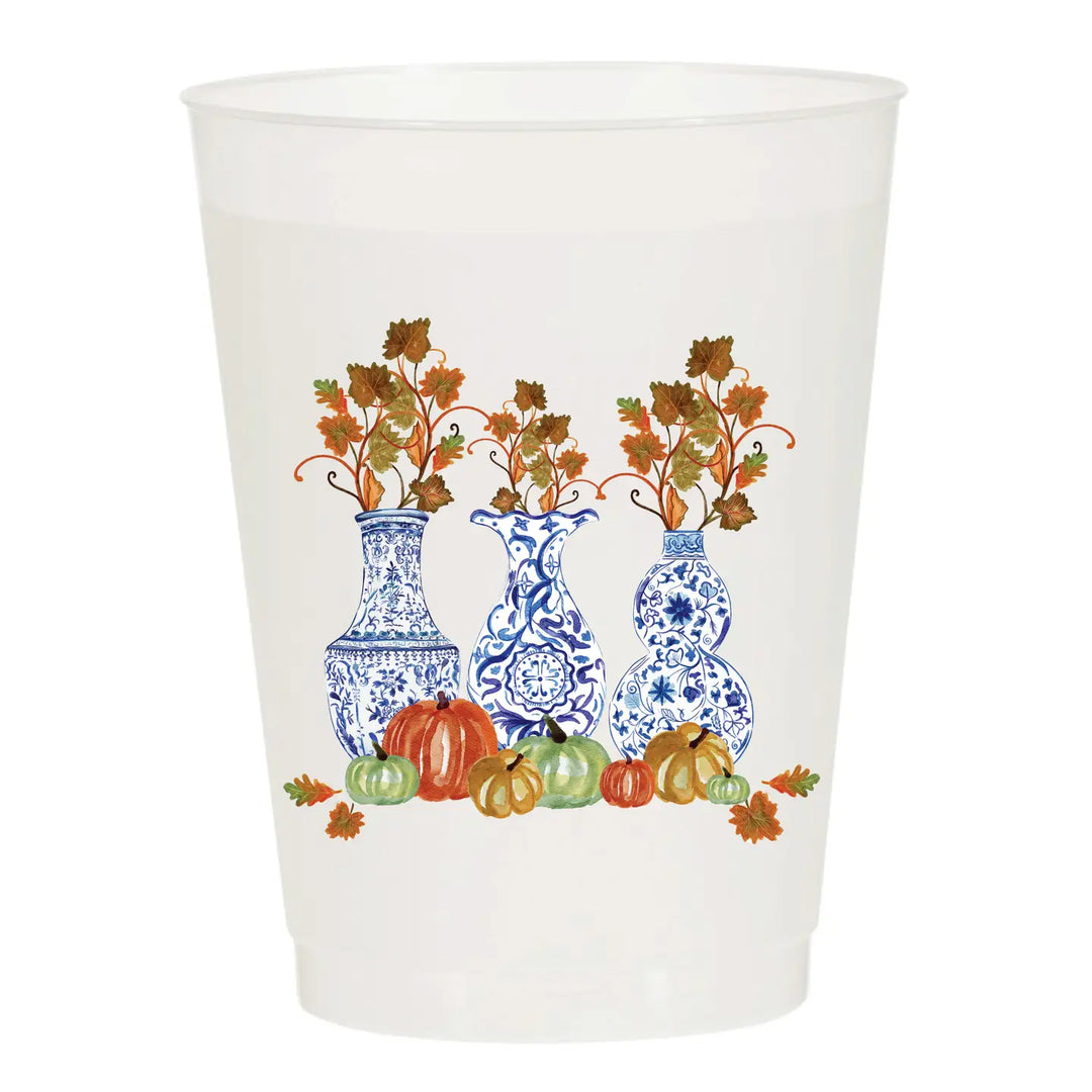 A white plastic cup featuring a printed autumn-themed design of three blue-and-white patterned vases filled with orange and brown leaves, surrounded by small pumpkins and fall leaves.
